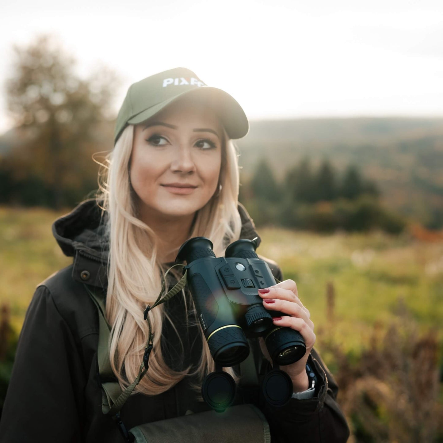 Close-up of a blonde model wearing a Pixfra branded hat holding the flagship Pixfra Draco D635-4K Multispectral Binoculars. The image demonstrates the ergonomic magnesium alloy chassis and highlights the large 35mm F1.0 thermal objective lens and 4K night vision sensor designed for professional thermal observation