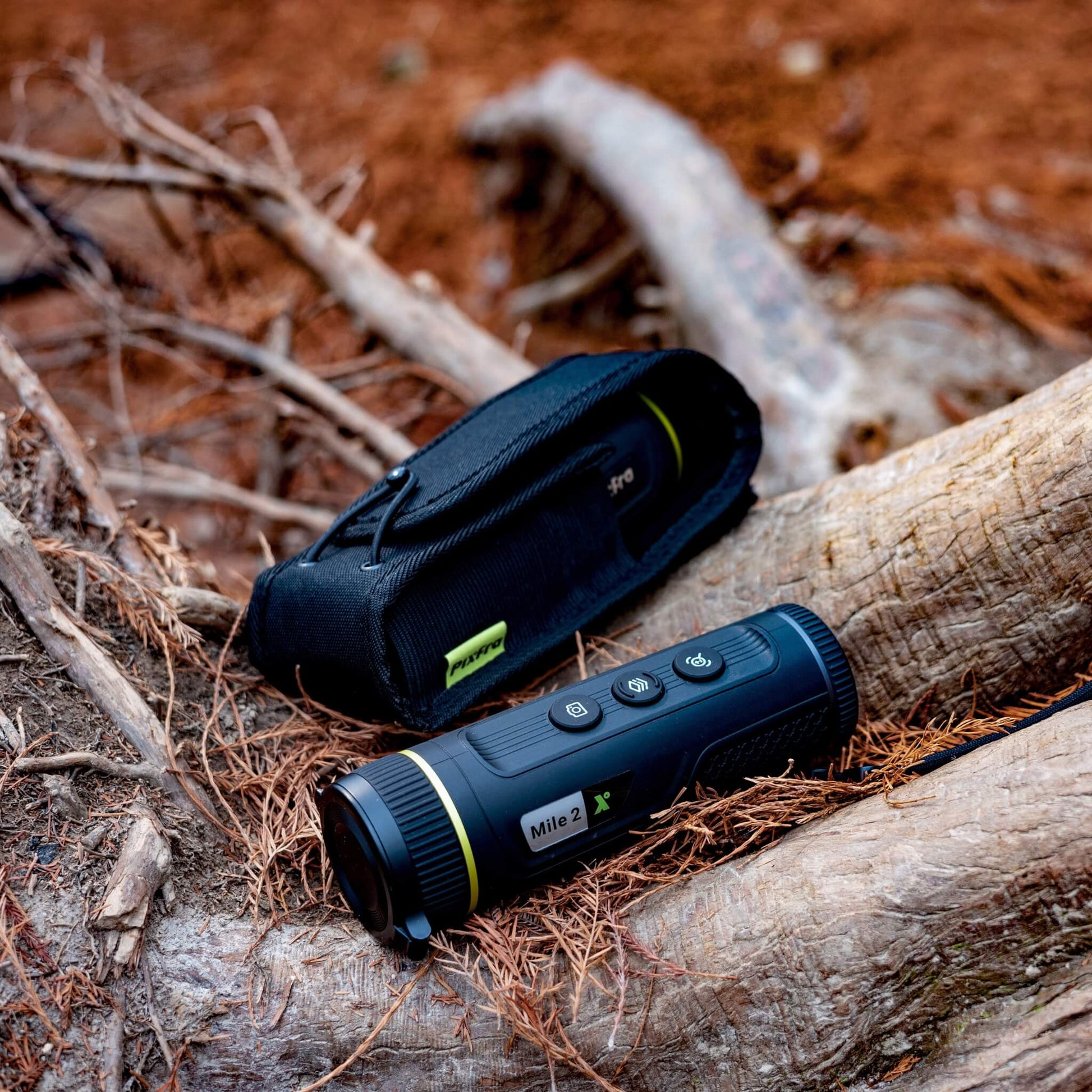 The Pixfra Mile 2 M425 Thermal Monocular sitting on a tree branch next to its protective carrying bag, emphasizing its portability and outdoor readiness.