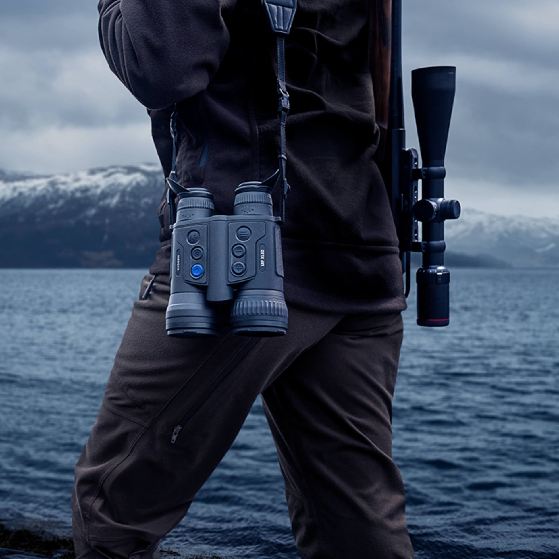 A person wearing hunting gear is carrying a pair of black Pulsar Merger LRF thermal binoculars on a strap over their shoulder. The binoculars are hanging in front of the person's right hip, facing outward with the lenses visible. The person is standing near a body of water with mountains in the background.