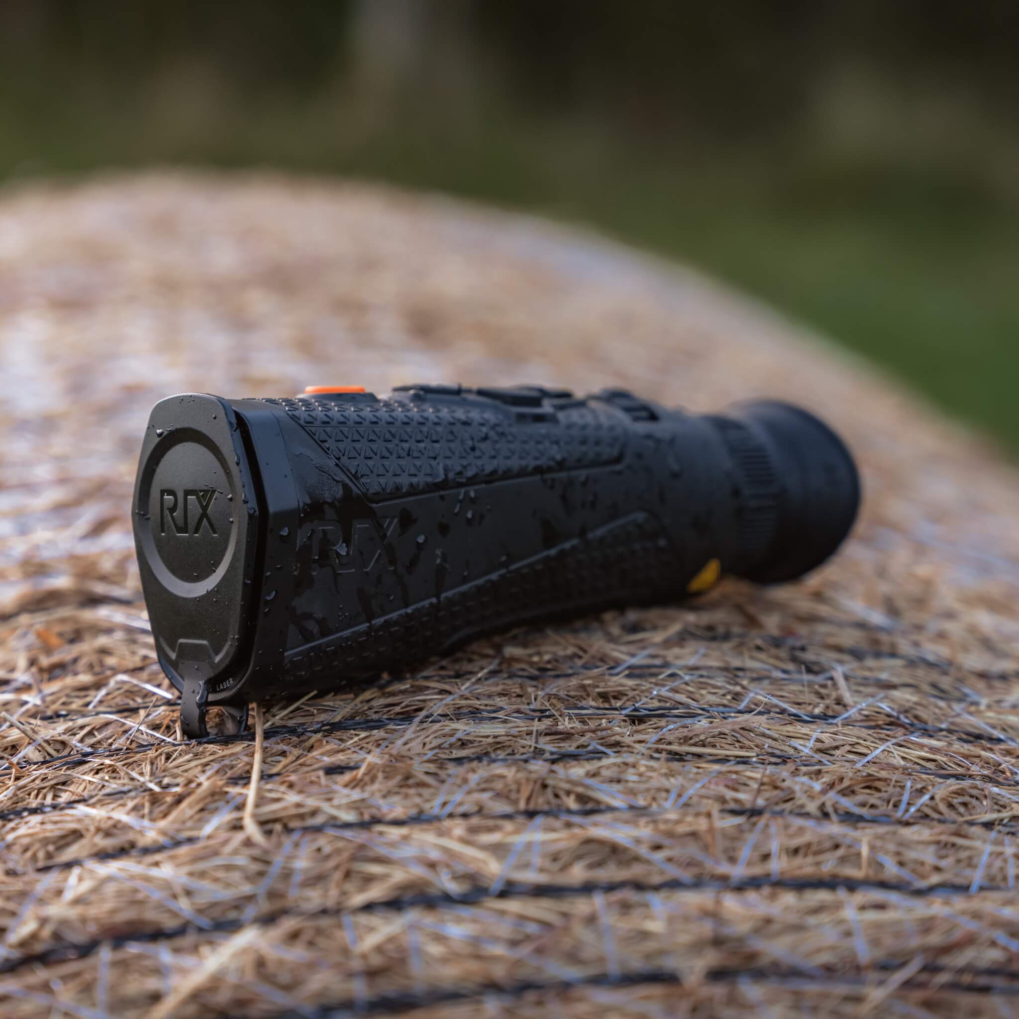 RIX Titan T3 Thermal Monocular sitting atop a hay bale with water droplets on the casing, showcasing its rugged IP67 waterproof and dustproof design for harsh outdoor environments.