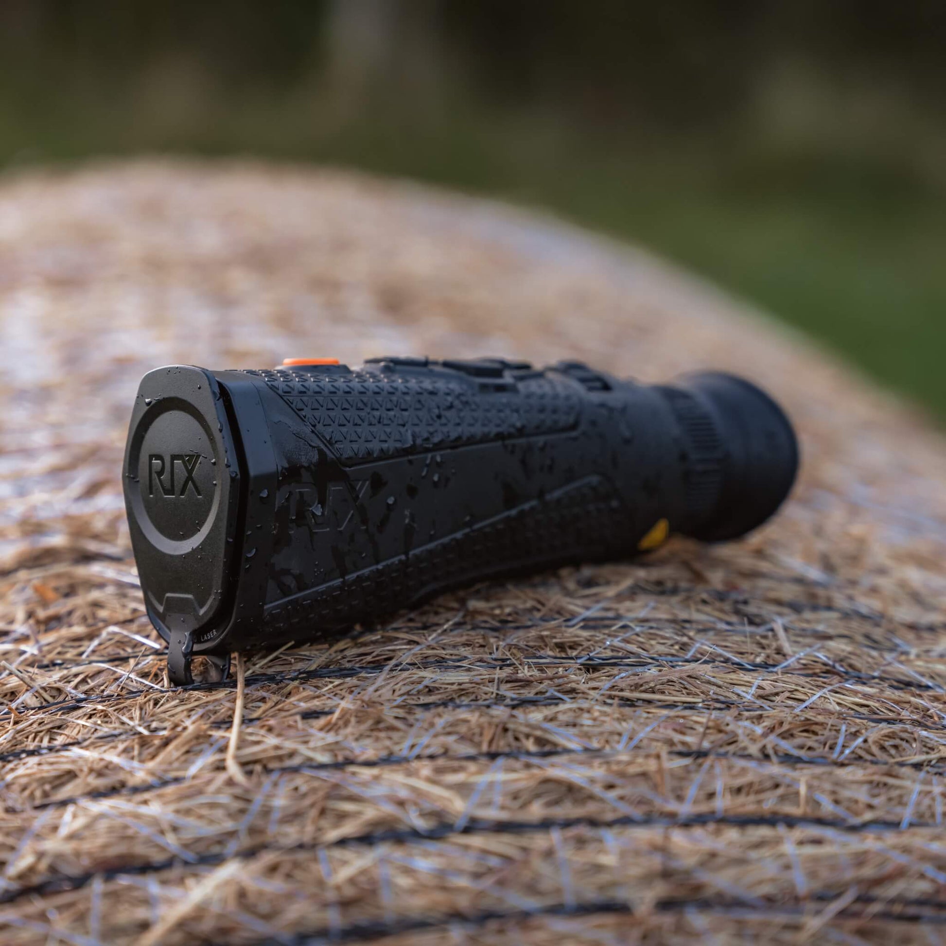 RIX Titan T3 Thermal Monocular sitting atop a hay bale with water droplets on the casing, showcasing its rugged IP67 waterproof and dustproof design for harsh outdoor environments.